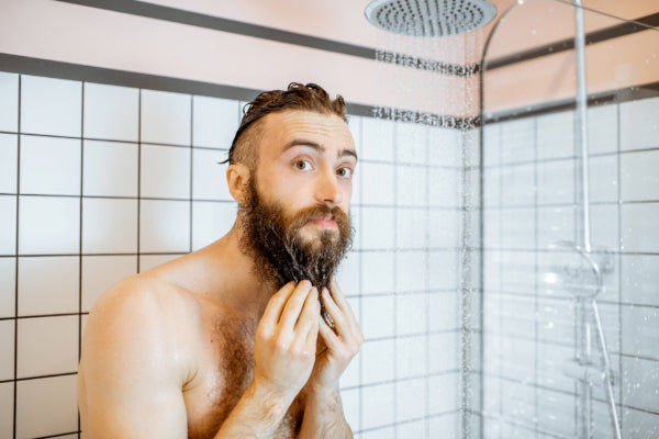 Man under running water in shower holding his beard looking at camera